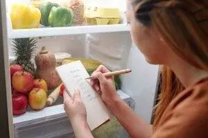Woman writing grocery list by fridge with fresh produce; Food shop prep