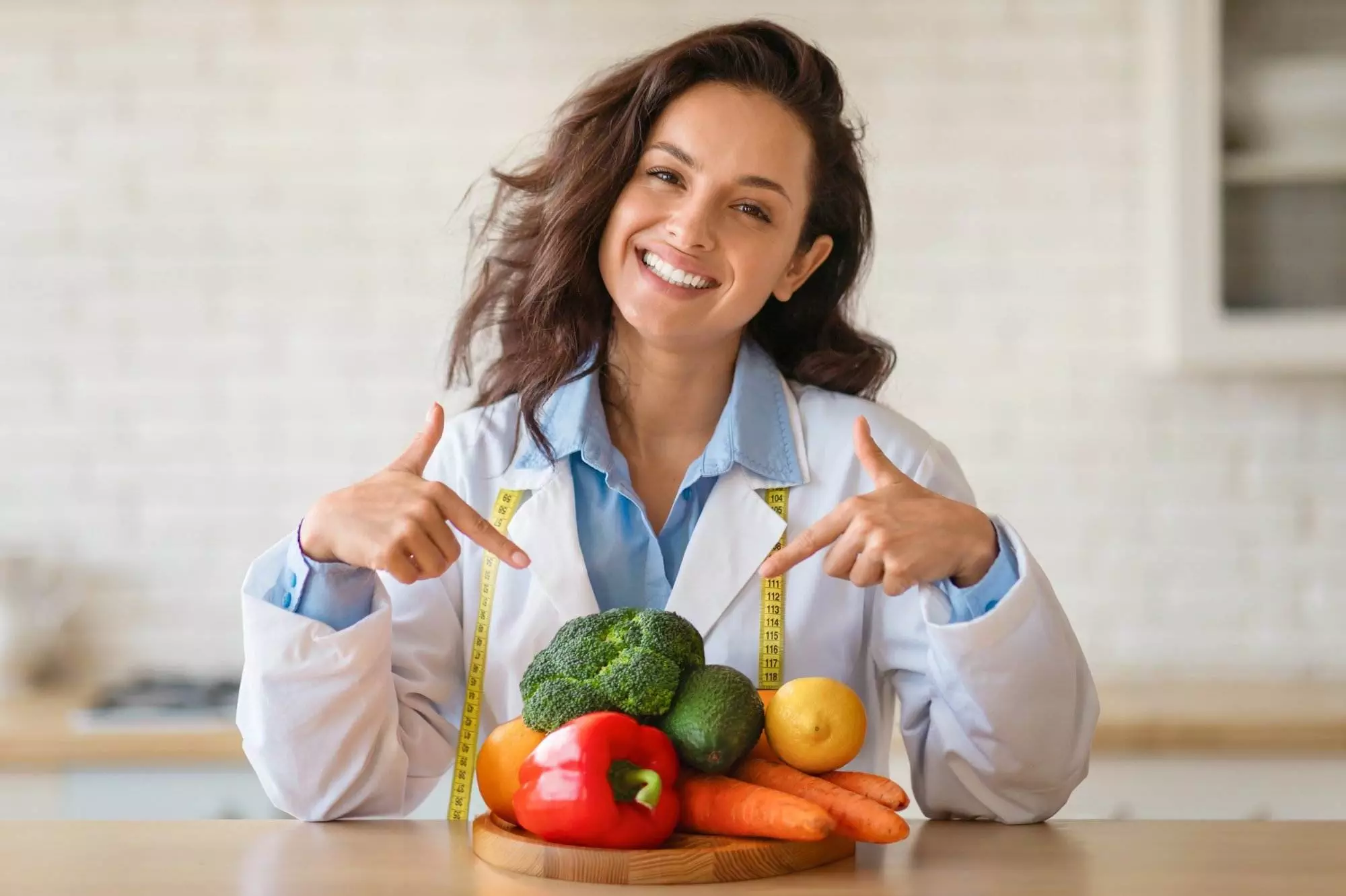 Joyful nutritionist in lab coat with tape measure draped around her neck, pointing enthusiastically at wooden board stacked with vibrant fruits and vegetables indoors