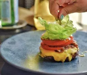 Hand adding lettuce to cheeseburger on plate.