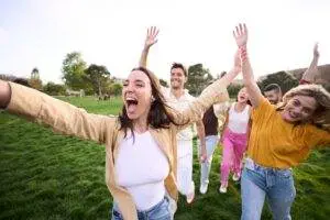 Group of six fit happy friends energized and raising arms in park after work, celebrating success.