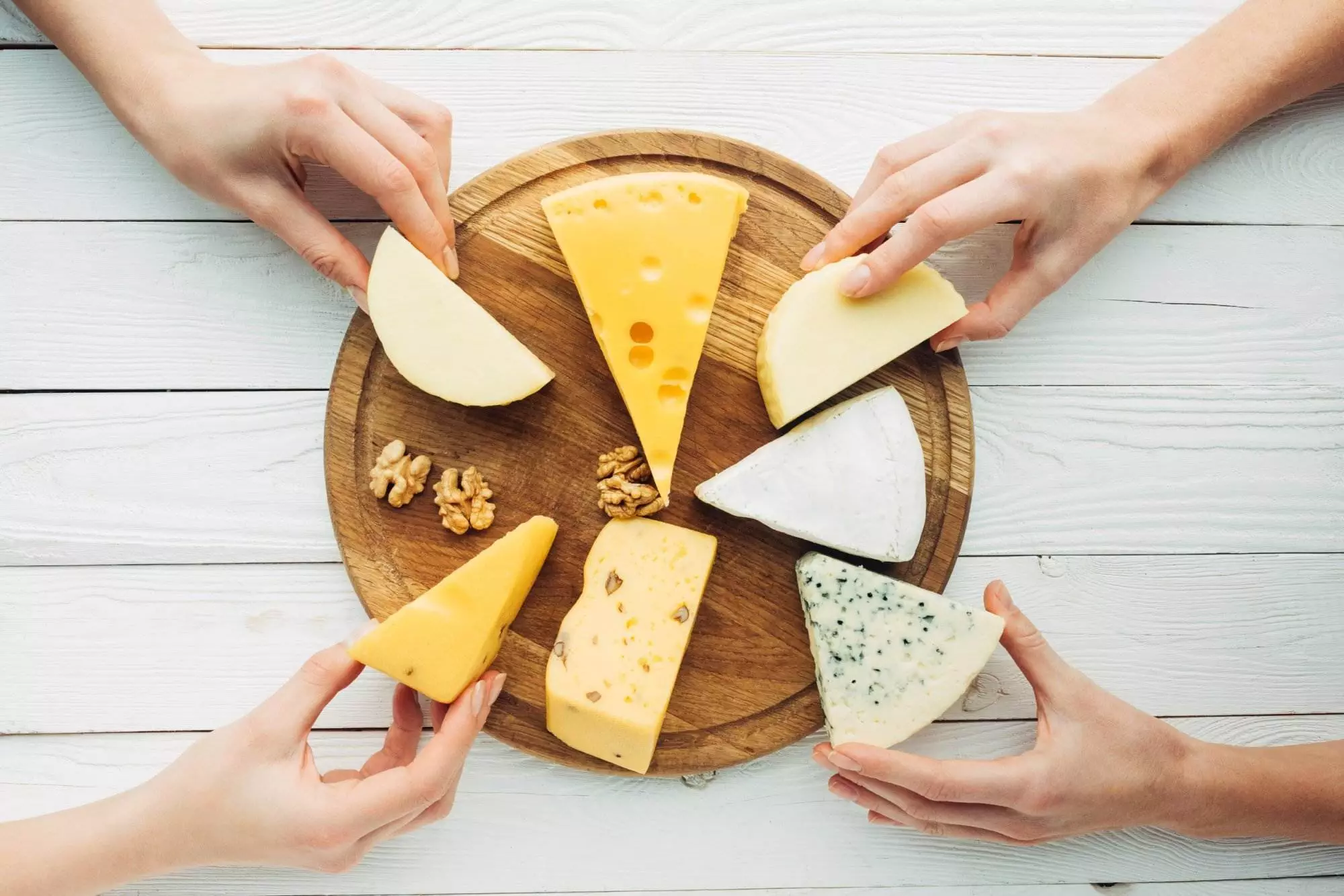 Hands selecting various cheeses on wooden board.