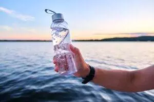 Hand holding water bottle by lake at sunset.
