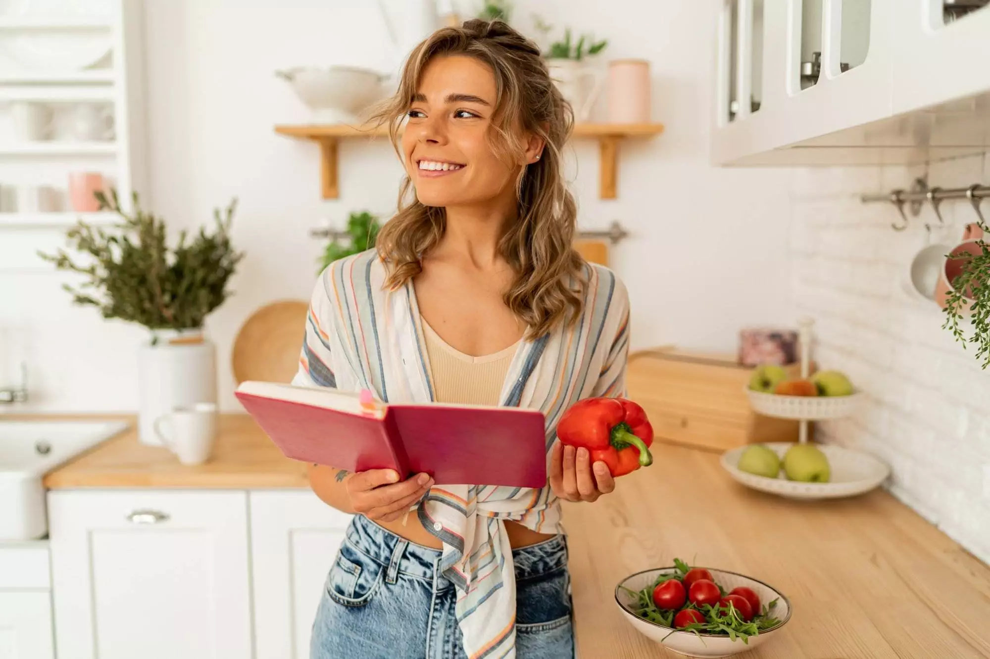 Woman with cookbook and bell pepper in kitchen.