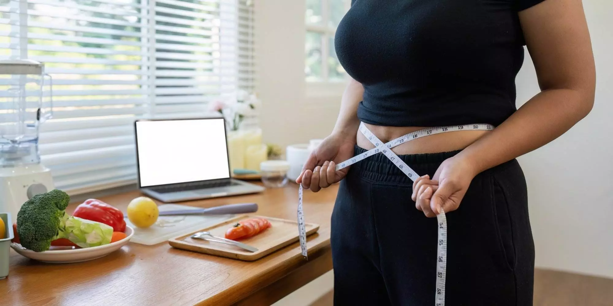 Person measuring waist in kitchen with healthy food.